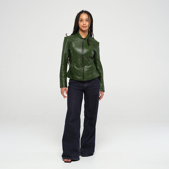 Woman standing against a plain studio background wearing a fitted green leather jacket and dark wide-leg jeans.
