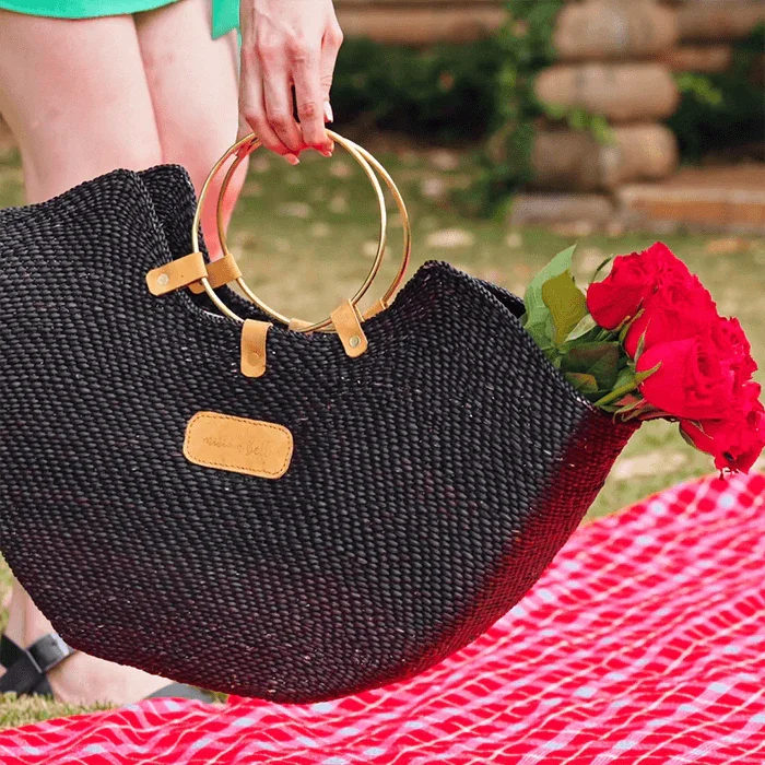 Zuri black handwoven seagrass tote bag with gleaming brass handles, holding red roses on a picnic blanket.