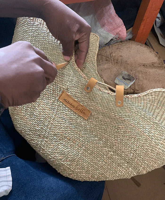 Artisan's hands attaching a leather "miriam bella" tag and strap to a natural straw seagrass tote bag.