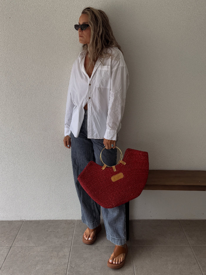 A woman stands against a plain beige wall wearing a white button-up shirt, blue jeans, and brown sandals, holding a red woven tote straw bag with a circular gold handle.