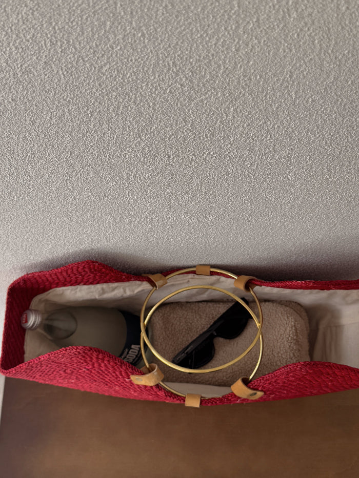A red woven tote bag with tan leather handles, shown from above on a neutral surface. Inside the bag are gold hoop earrings, black sunglasses, and a beige textured pouch.