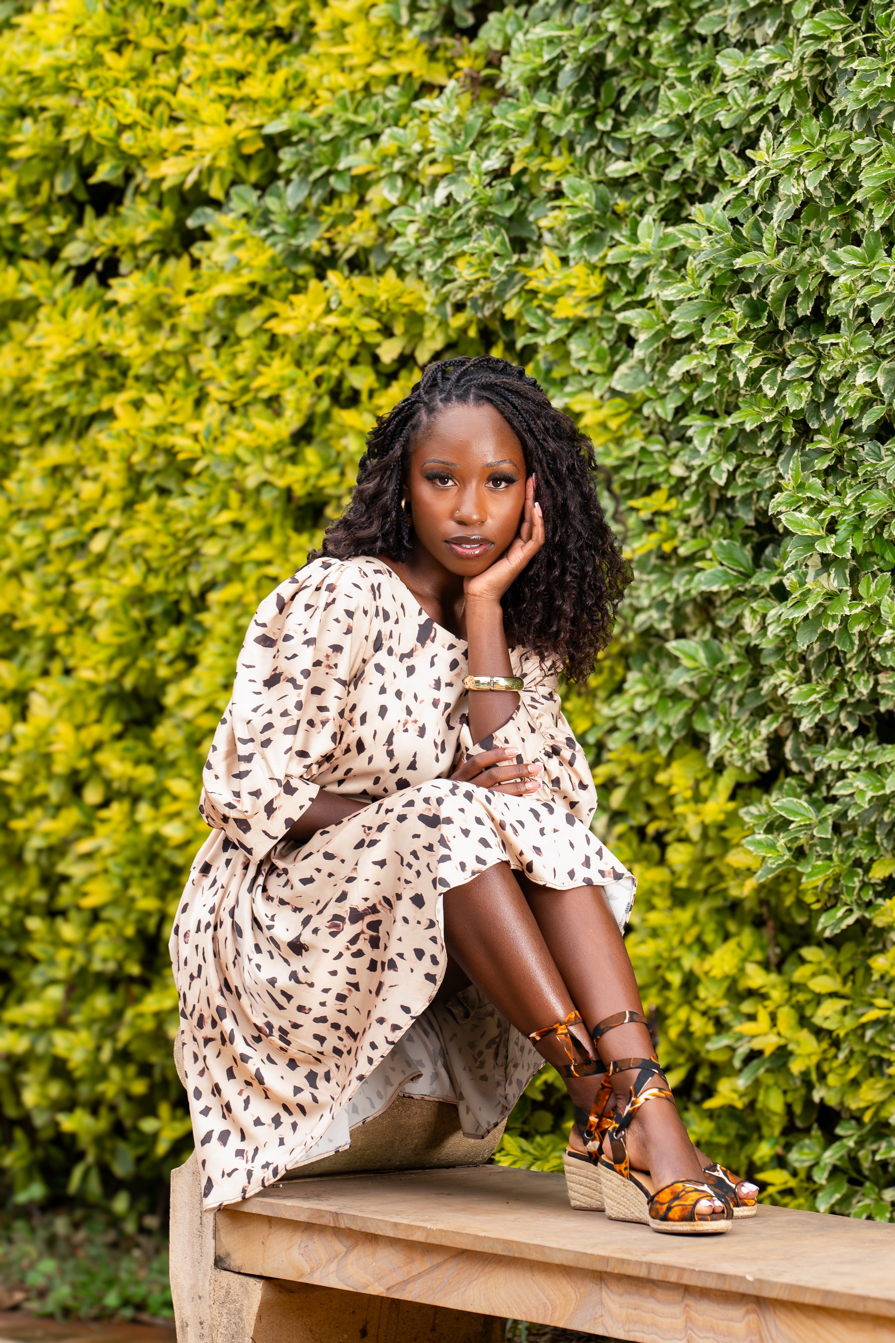 Woman in a patterned dress and limited edition espadrille wedg sandals sitting on a wooden bench with greenery in the background