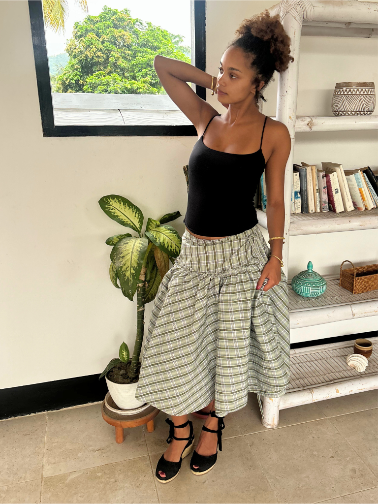Woman in black top and plaid skirt standing in a room with a plant and bookshelf.