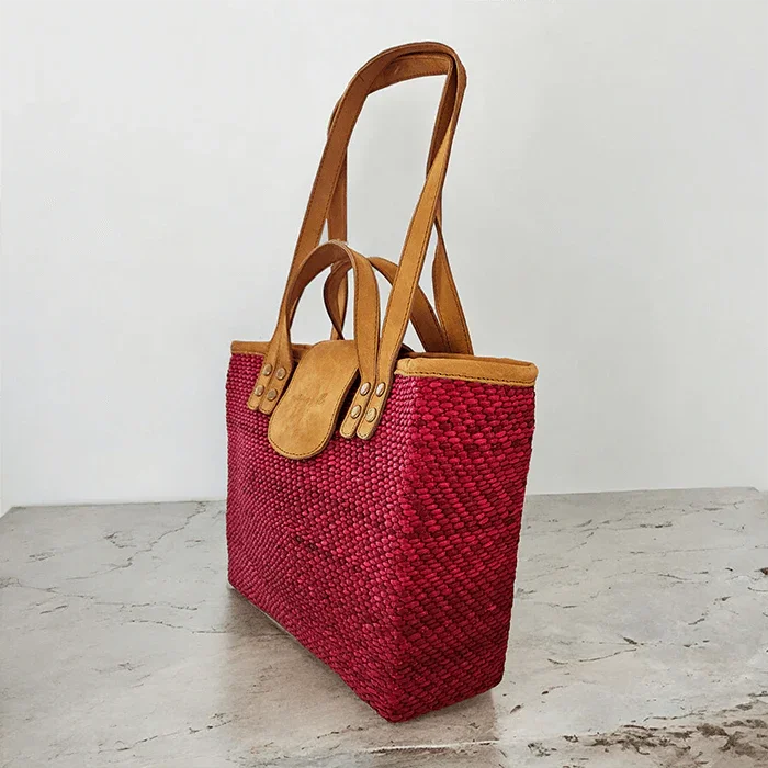 Red woven tote bag with brown leather handles on a light wooden surface.