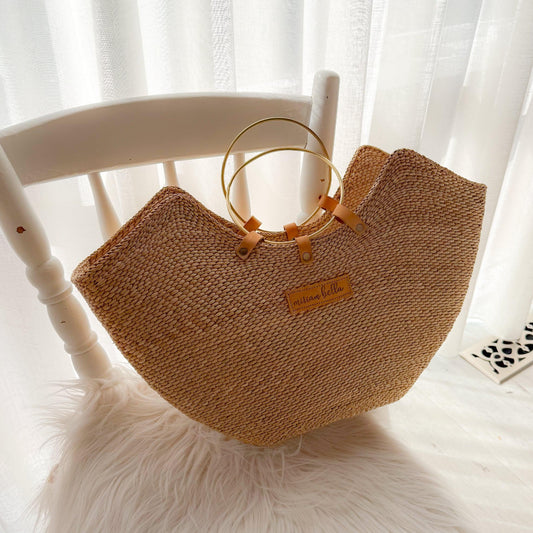 A natural woven straw tote bag with a semi-circular shape, featuring a small tan leather patch with the brand name and two gold-tone circular handles, resting on a white faux fur rug in front of a white wooden chair.