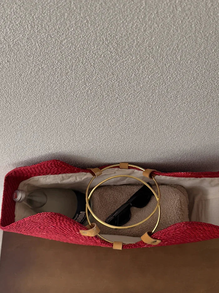 Red woven straw beach bag with brass handles, holding a water bottle, towel, and sunglasses.