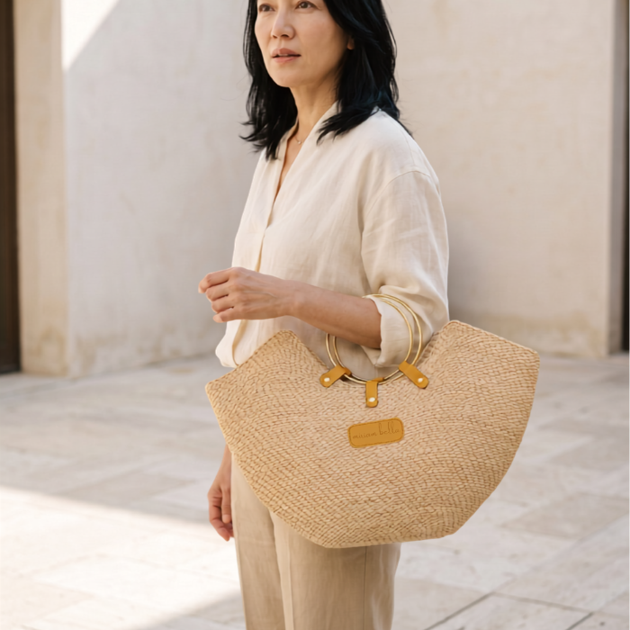 A woman in a cream outfit holding a natural coloured Jua woven straw beach bag in with brass ring handles.