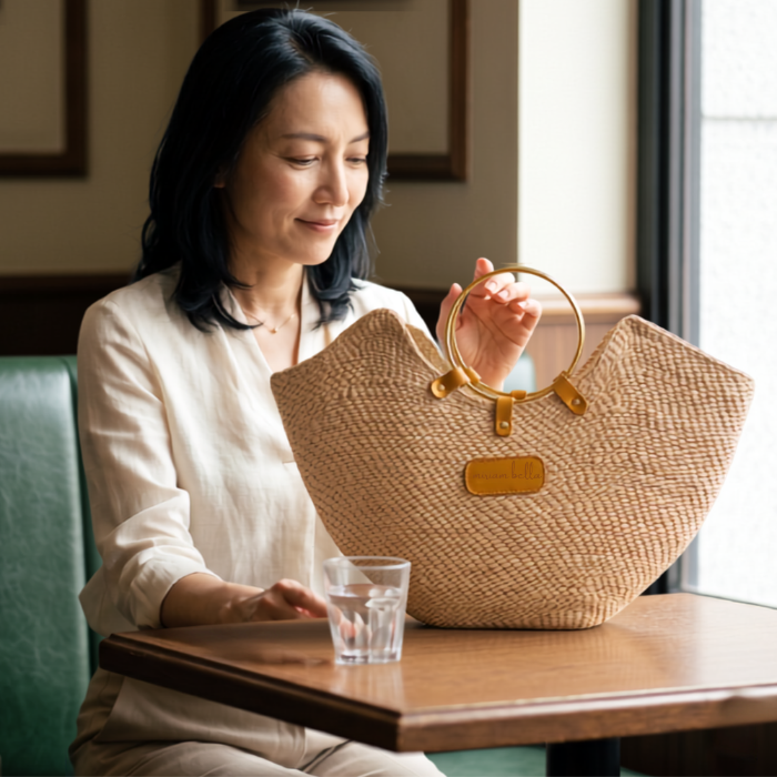 A woman in a cafe sitting next to natural coloured Jua woven straw beach bag with brass ring handles in black.