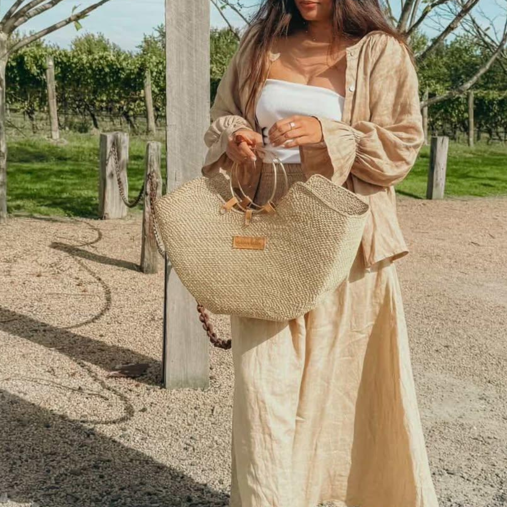 A woman outdoors wearing a beige linen outfit and holding a large woven straw tote bag with a brown leather detail.