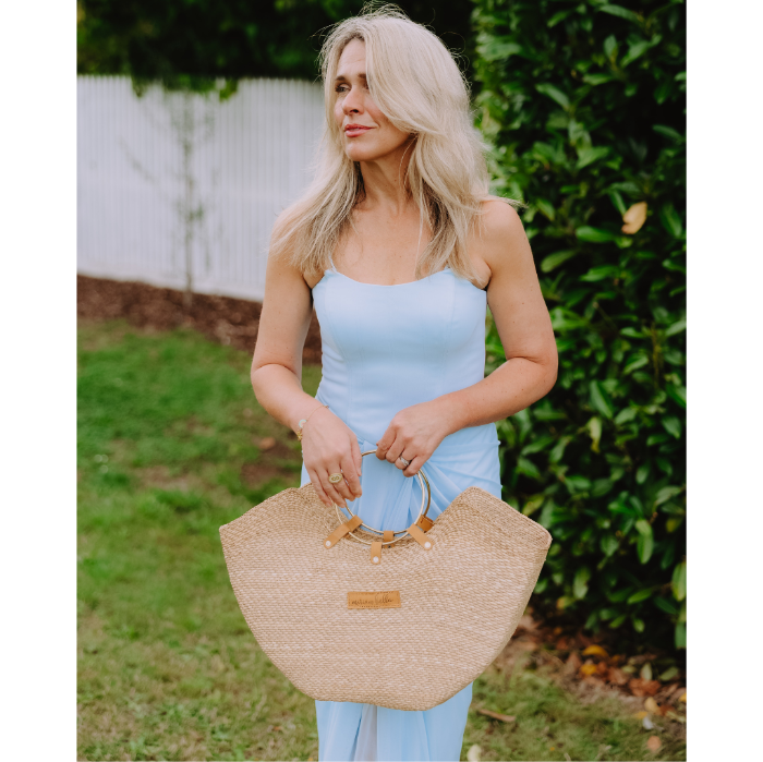 Woman in a blue dress holding a Jua woven straw beach bag with brass ring handles.