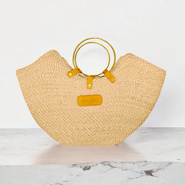Handcrafted Jua woven straw beach bag with brass ring handles and a tan leather logo patch.