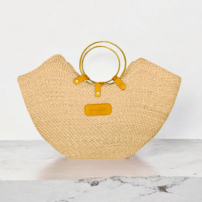 Handcrafted Jua woven straw beach bag with brass ring handles and a tan leather logo patch.