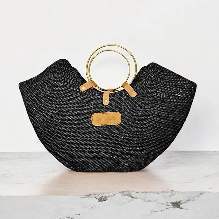 Black woven straw beach bag with elegant brass ring handles and a tan leather label.
