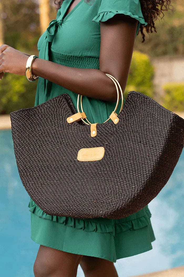 Black woven straw beach bag with brass ring handles and a tan leather tag, held by a woman in a green dress.