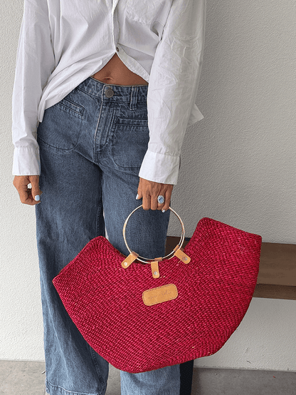 Red woven straw beach bag with brass handles, held by a person in jeans and a white shirt.