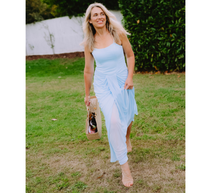 Blonde woman in a light blue dress carrying a handcrafted woven straw beach bag with brass handles.