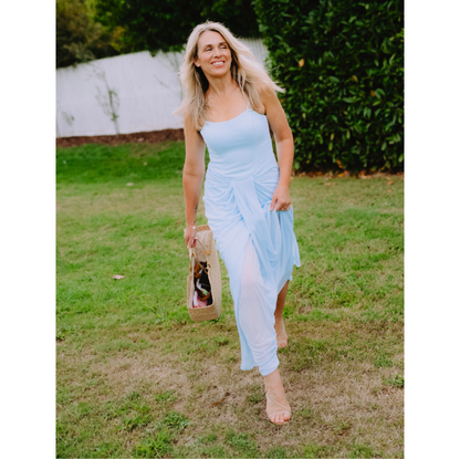 Blonde woman in a light blue dress carrying a handcrafted woven straw beach bag with brass handles.