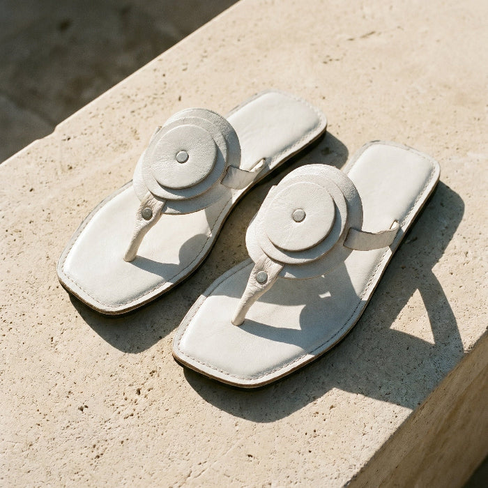 Pair of white leather flat sandals with a unique circle detail and thong strapon a stone surface