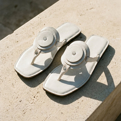 Pair of white leather flat sandals with a unique circle detail and thong strapon a stone surface