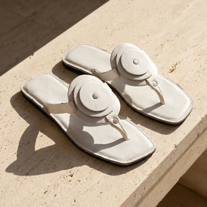 Pair of Women's white leather flat sandals with a unique circle detail and thong strap on a light concrete surface