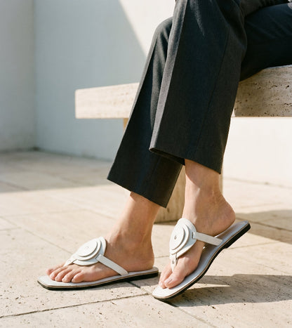 Person wearing a pair of women's white leather flat sandals with a unique circle detail and thong strap with black soles on a light concrete floor.