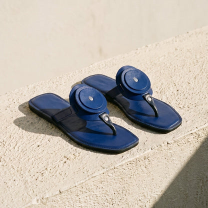 Pair of Women's blue leather flat sandals with a unique circle detail and thong strap on a light stone surface.