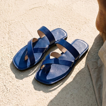 A pair of handmade indigo leather sandals with cross strap produced by Australian brand Miriam Bella on a sandy surface with a person wearing beige pants.