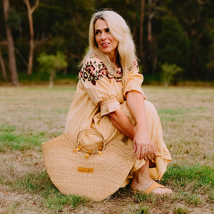 Woman in a yellow dress wearing tan women's leather slides sitting on grass with a straw bag