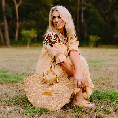 Woman in a yellow dress wearing tan women's leather slides sitting on grass with a straw bag
