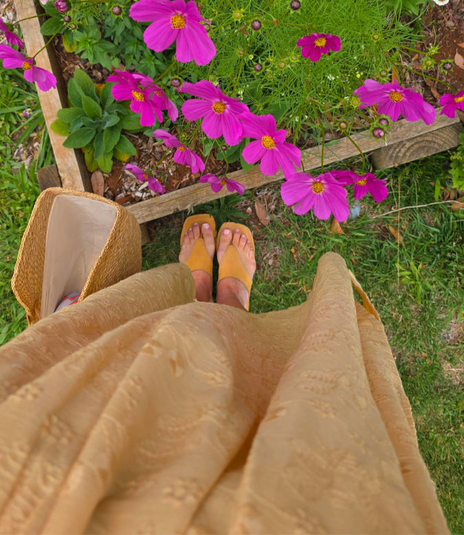 Person wearing a beige dress and tan Women's leather slide sandals standing on grass with pink flowers in the background.