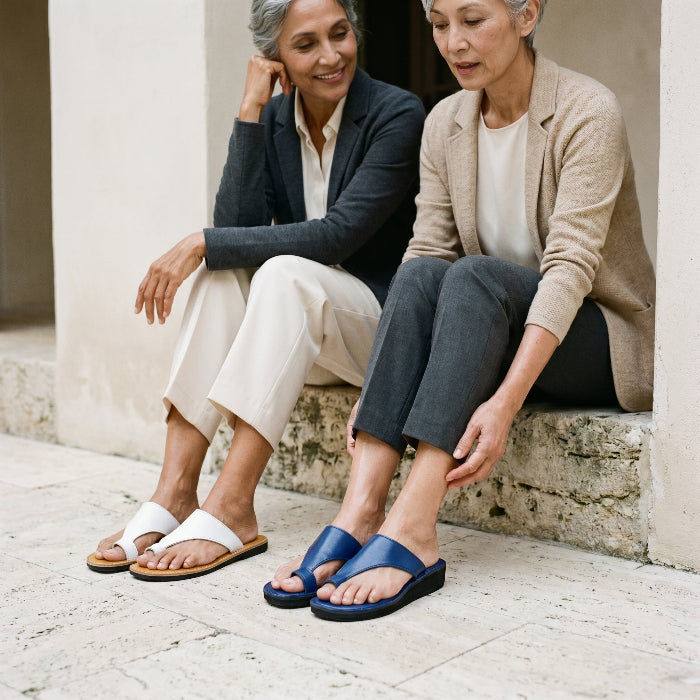 Two women sitting on a stone ledge thong wearing sandals by Miriam Bella.