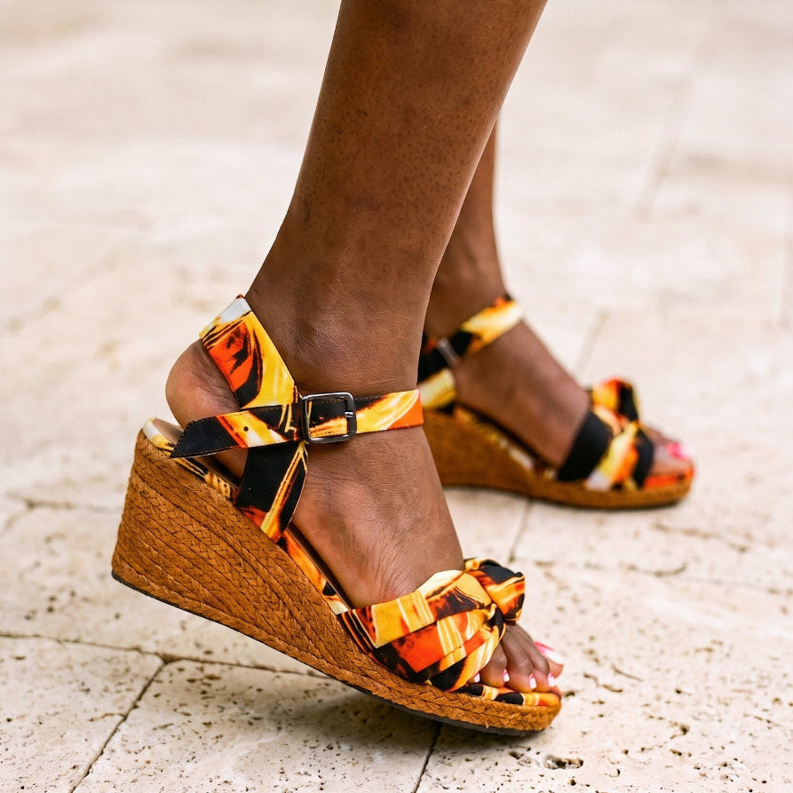 A person wearing wedge sandals with a bold orange, yellow, and black patterned fabric upper and woven jute wedge soles, standing on a light stone pavement.