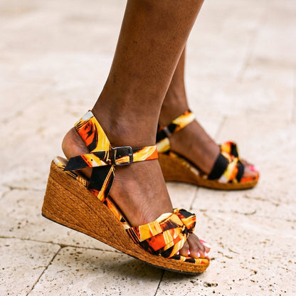 A person wearing wedge sandals with a bold orange, yellow, and black patterned fabric upper and woven jute wedge soles, standing on a light stone pavement.