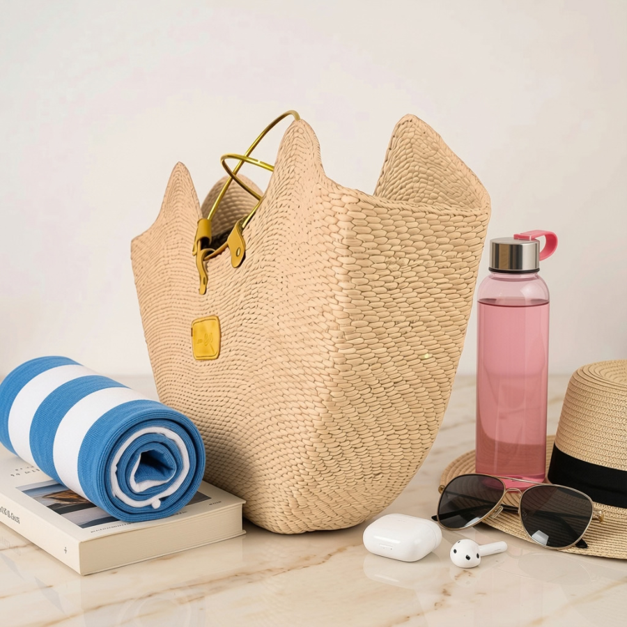 The Jua large woven straw tote bag with gold-tone handles and a small gold patch, styled on a light marble surface with a blue-and-white striped towel, pink water bottle, sunglasses, white earbuds case, and a straw hat.