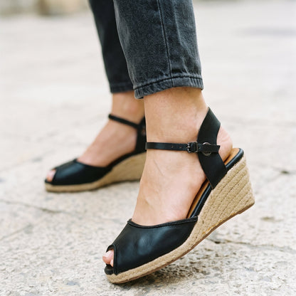 A person wearing black peep-toe wedge sandals with espadrille-style woven wedge heels, paired with dark gray jeans, standing on a light stone pavement.