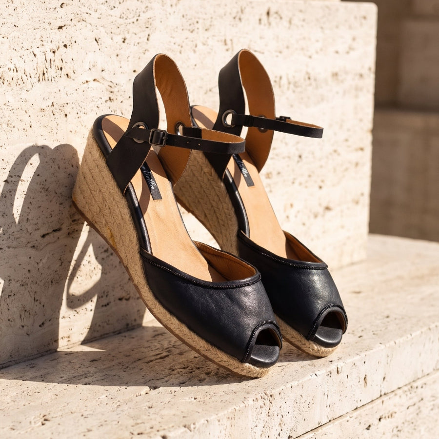 A pair of black leather wedge sandals with open toes and ankle straps, resting on light beige stone steps outdoors.
