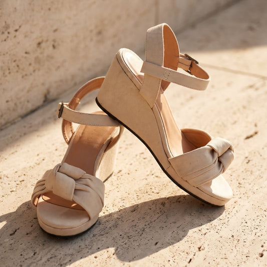A pair of beige wedge sandals with ankle straps and a knotted front detail, photographed on a sunlit stone surface against a textured wall.