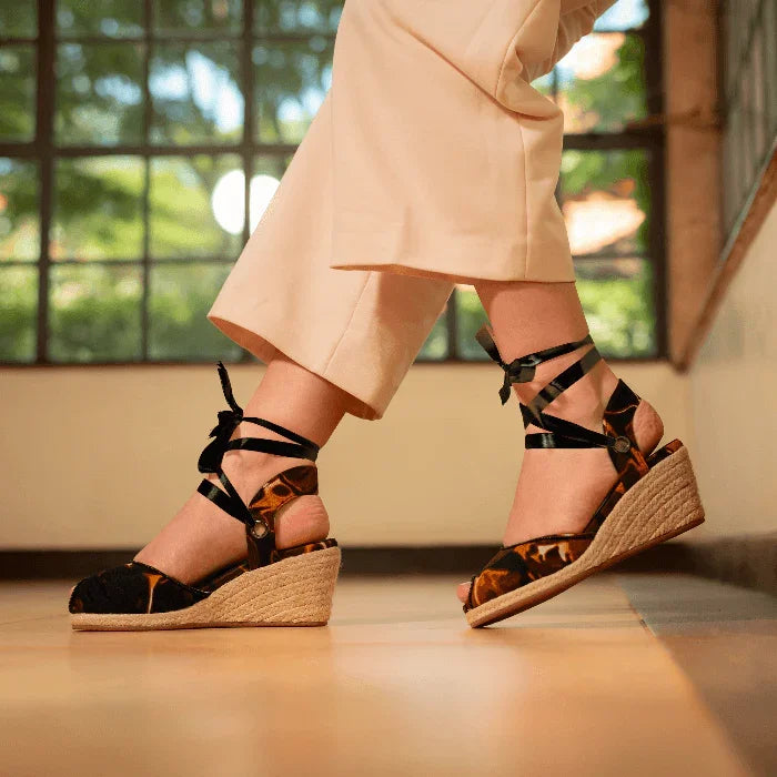 Woman wearing Gaia Midnight Flame espadrille wedge heels with black ankle ties and cream trousersbackground.