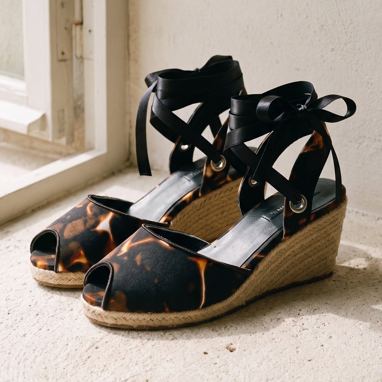 A pair of wedge sandals with black lace-up ankle straps and a black-and-brown mottled peep-toe upper, resting on a sunlit stone ledge next to a window.