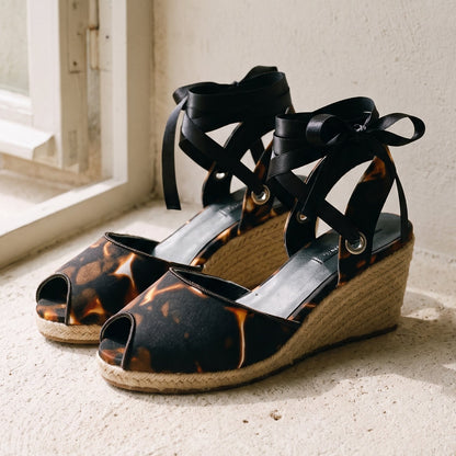 A pair of wedge sandals with black lace-up ankle straps and a black-and-brown mottled peep-toe upper, resting on a sunlit stone ledge next to a window.