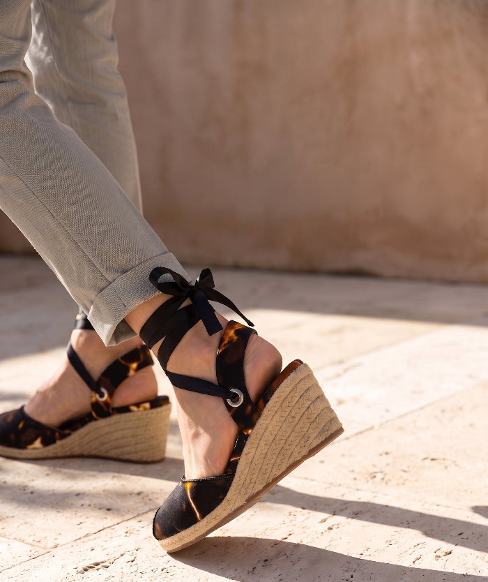 Woman wearing Gaia Midnight Flame espadrille wedge heels with black ankle ties and blue trousers.