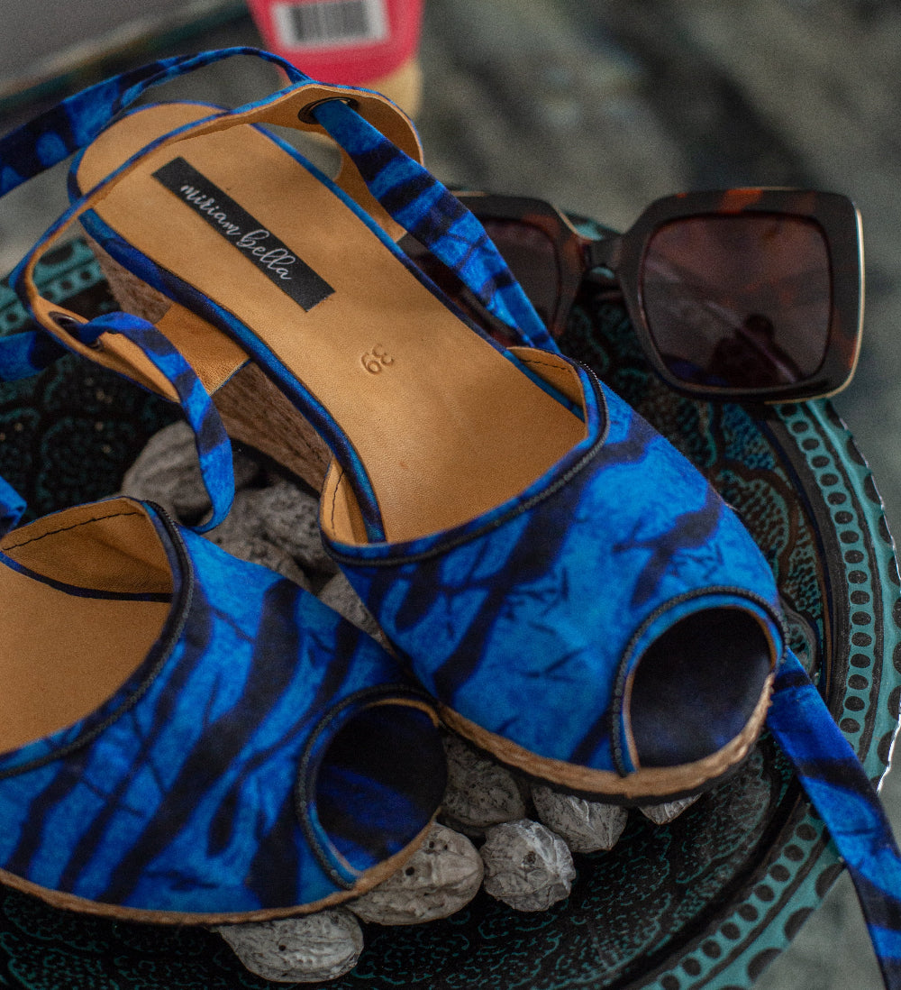 Blue and black patterned peep-toe heeled wedge sandals with cork-look heels, displayed on a decorative tray with sunglasses in the background.