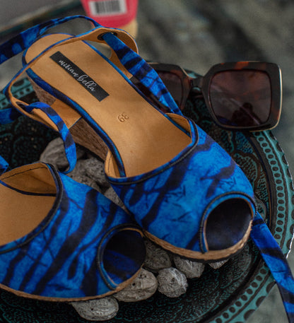 Blue and black patterned peep-toe heeled wedge sandals with cork-look heels, displayed on a decorative tray with sunglasses in the background.