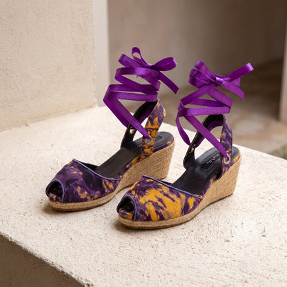 Pair of Gaia Royal Ember espadrille wedge heels with purple ankle ties and peep toe design on a stone bench.