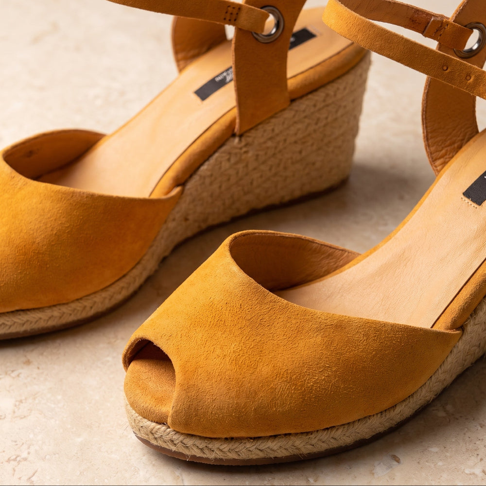  A pair of handcrafted leather ochre wedge espadrille sandals with jute sole by Miriam Bella with adjustable ankle straps and braided jute wedge heels,  on a light beige textured surface.