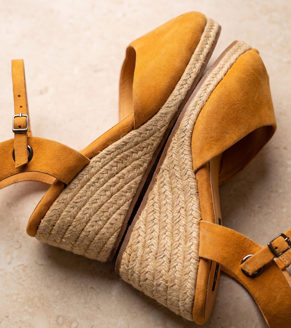 A pair of handcrafted leather ochre wedge espadrille sandals with jute sole by Miriam Bella with adjustable ankle straps and braided jute wedge heels, displayed on a light beige textured surface.