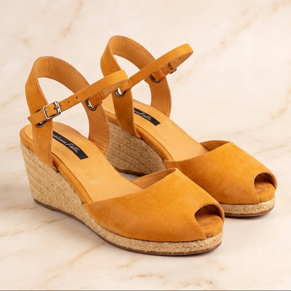 A pair of handcrafted leather ochre wedge espadrille sandals with jute sole by Miriam Bella with adjustable ankle straps and braided jute wedge heels, displayed on a light beige marble surface.