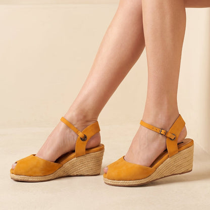 A pair of handcrafted leather ochre wedge espadrille sandals with jute sole and ankle straps by Miriam Bella, shown on a model's feet against a warm beige background.