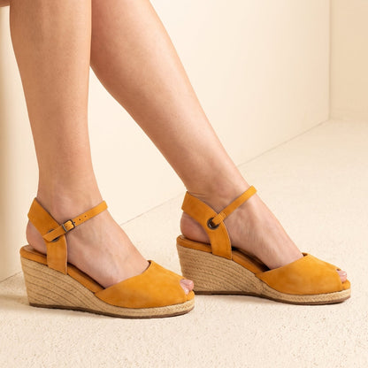 A pair of handcrafted leather ochre wedge espadrille sandals with jute sole and ankle straps by Miriam Bella, shown on a model's feet against a warm beige background.