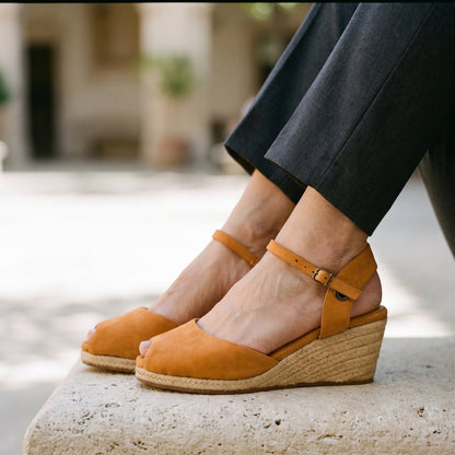 A person wearing tan leather wedge espadrille sandals with ankle straps and dark cropped trousers, seated on a stone bench in an outdoor setting.